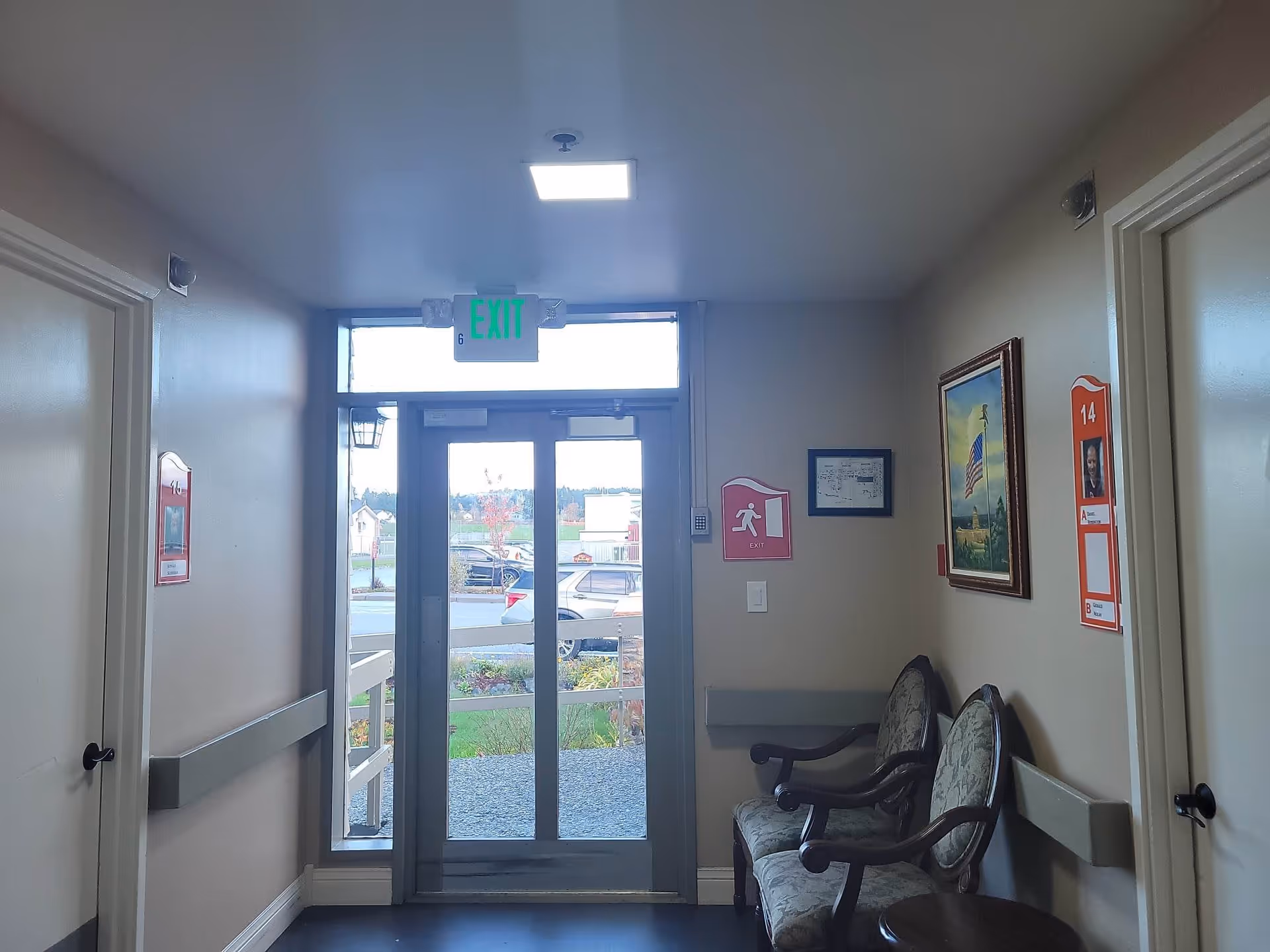 Interior hallway of a senior living facility with an exit door leading outside. The hallway has beige walls, two cushioned chairs with wooden arms, a small round table, and framed pictures on the walls. There are room number signs on the doors and an illuminated green exit sign above the door.