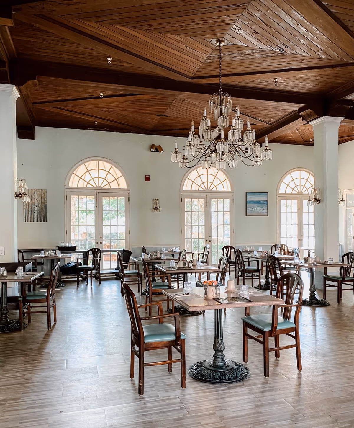 Sunlit dining room with wooden tables and chairs, a decorative chandelier, and arched French doors and windows.