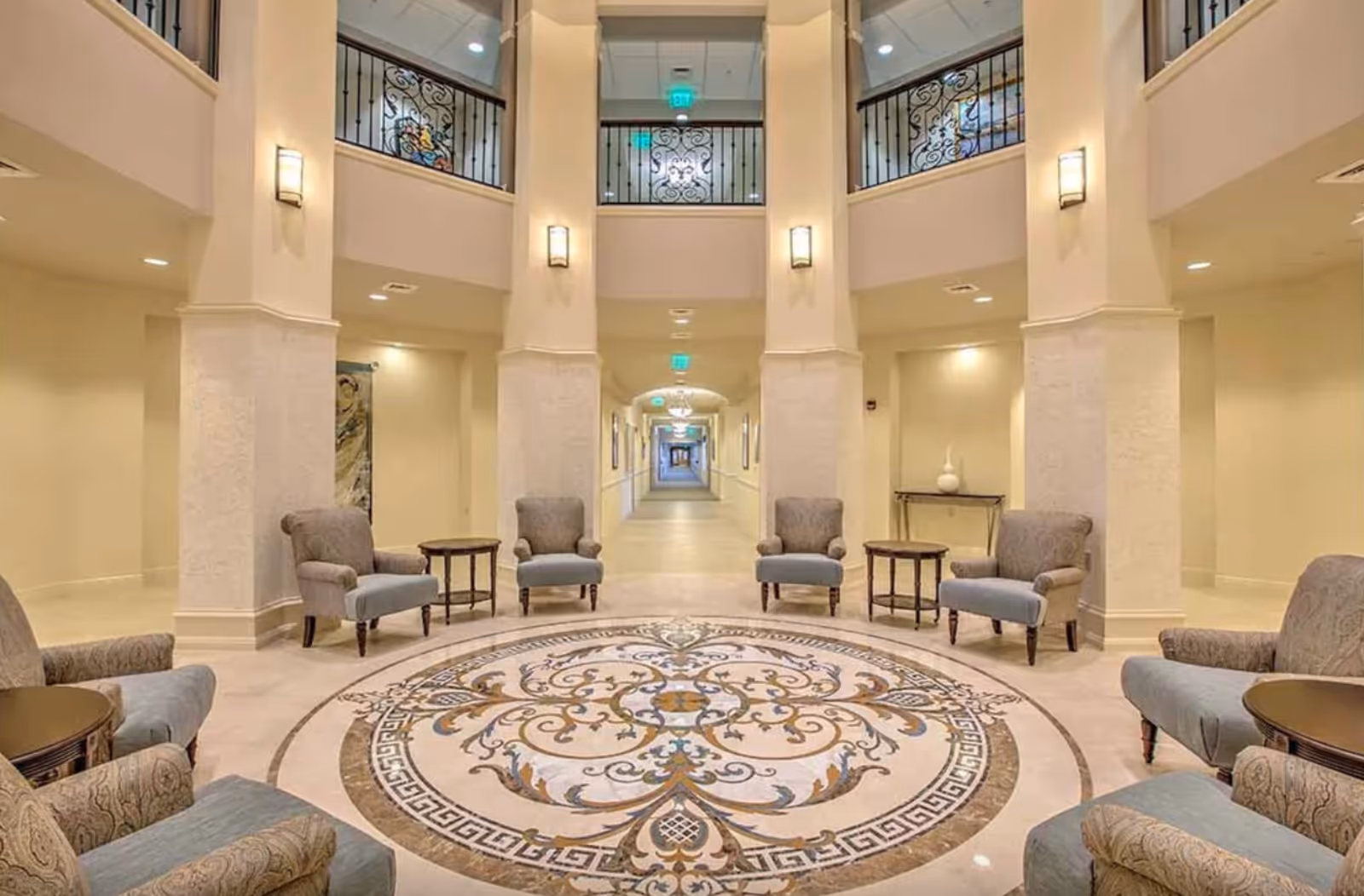 Spacious elegant interior lobby with a circular decorative floor medallion, armchairs and side tables arranged around it and a long corridor beyond.