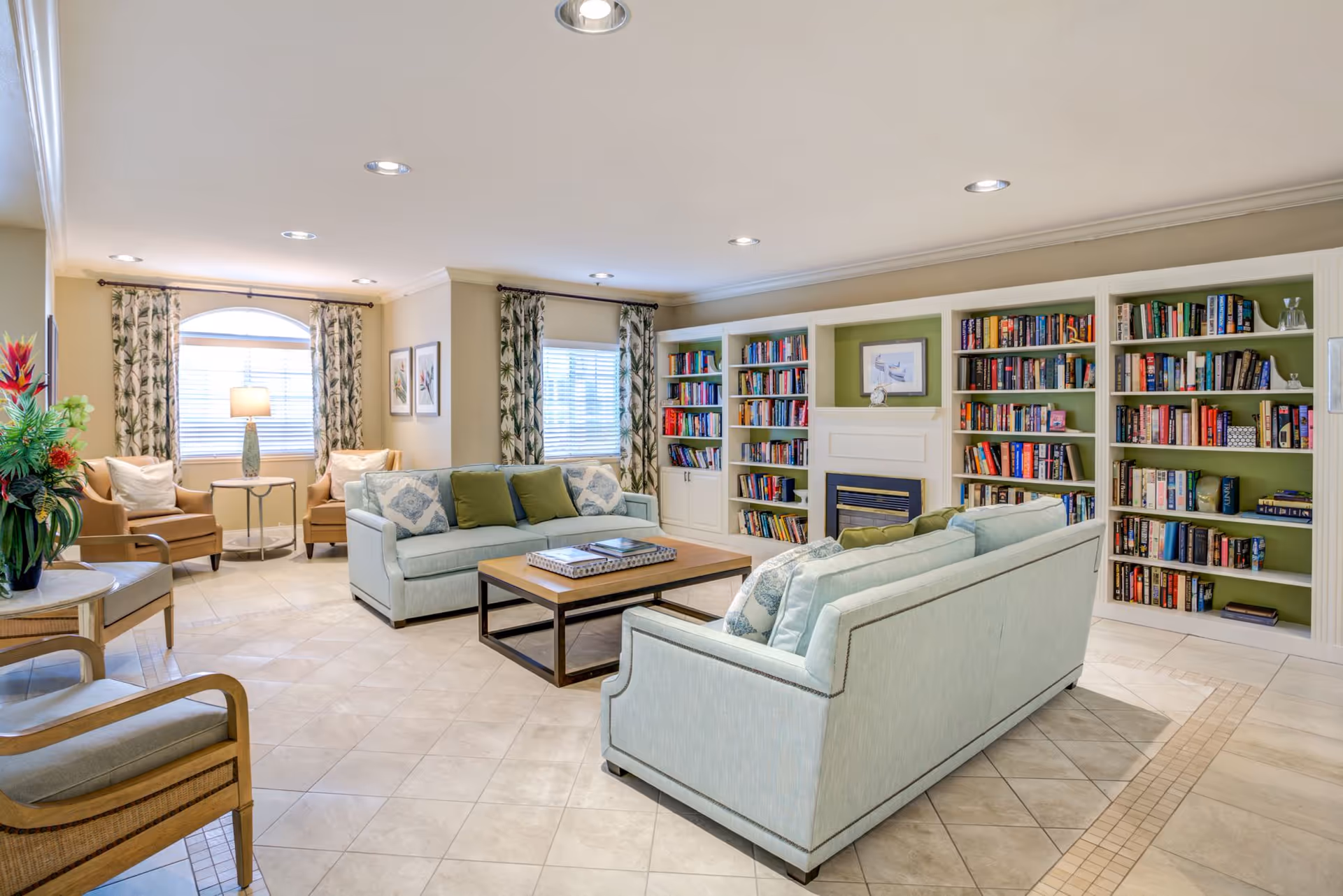 A bright and spacious living room with two light blue sofas facing each other around a wooden coffee table. There are built-in bookshelves filled with books and a fireplace in the center. The room has tiled floors, recessed ceiling lights, two windows with patterned curtains, and additional seating with armchairs and a side table with a lamp.