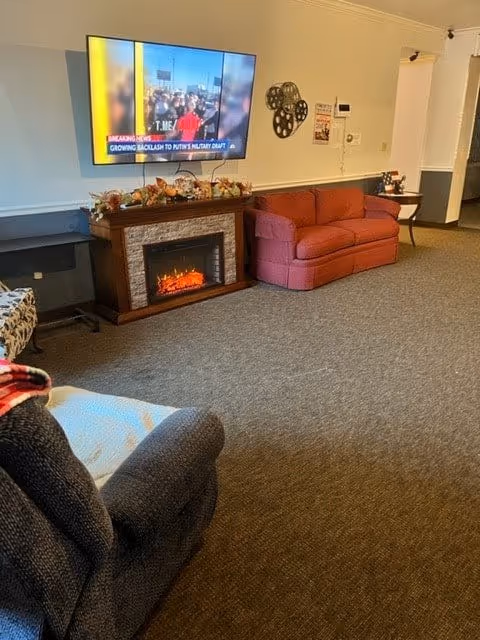 A cozy living room area with a mounted flat-screen TV displaying a news channel. Below the TV is an electric fireplace decorated with autumn-themed garlands. To the right of the fireplace is a red loveseat, and in the foreground, there is a dark upholstered armchair with a blanket draped over one arm. The room has beige walls and carpeted flooring.