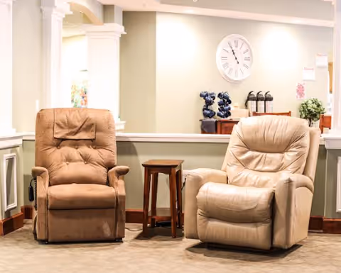 Two cushioned recliner chairs, one brown and one beige, positioned side by side in a well-lit room with a small wooden side table between them. In the background, there is a wall clock, a wine rack with bottles, and some fire extinguishers mounted on the wall.