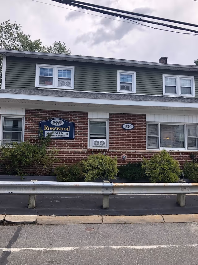 Exterior front view of a two-story assisted living facility named Rosewood Homestyle Assisted. The building has a brick lower half and green siding on the upper half, with several windows and bushes in front. A metal guardrail runs along the edge of the sidewalk in front of the building.