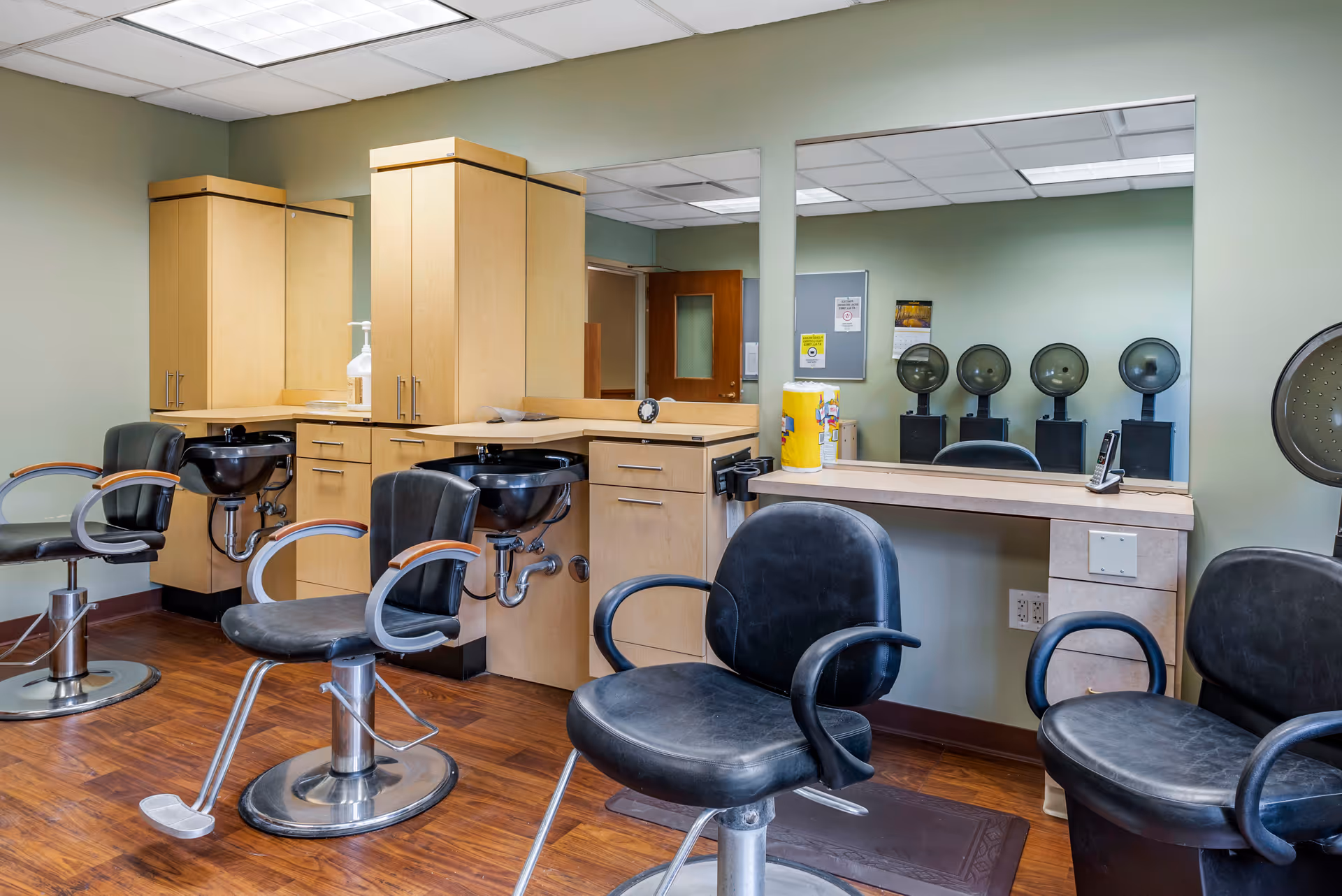 Interior view of a salon area with black salon chairs, wooden cabinetry, sinks for hair washing, large mirrors on the walls, and hair drying stations in the background.
