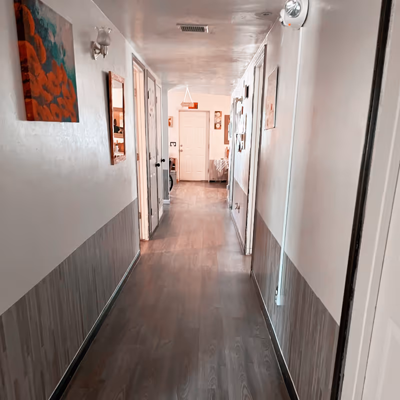 A long indoor hallway with wooden flooring and light-colored walls. Several doors line the hallway on both sides, with artwork and a mirror hanging on the left wall. At the end of the hallway, there is a closed white door and some furniture visible in the adjacent room.
