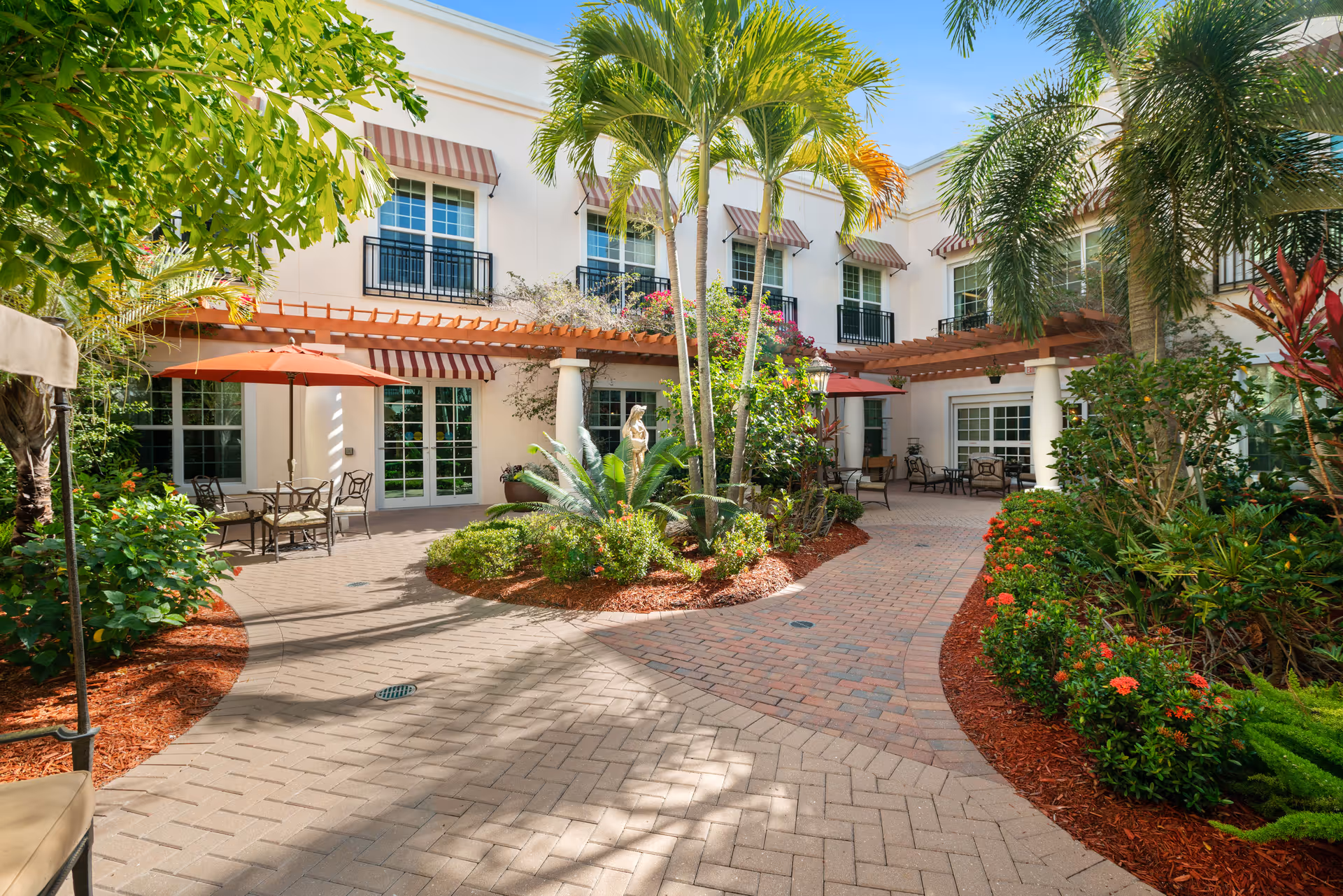 Outdoor courtyard area at Villa at Terracina Grand featuring a paved walkway, lush greenery, palm trees, flowering plants, and patio seating with umbrellas. The building has white walls with striped awnings over the windows and doors.