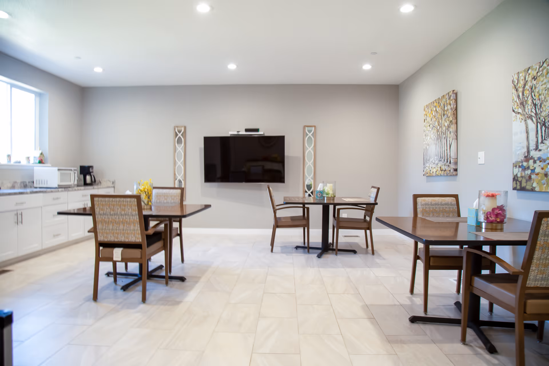 A bright dining room with three wooden tables, each surrounded by chairs with woven backs. The room has light-colored tiled flooring, neutral gray walls, and two paintings of trees on the right wall. A flat-screen TV is mounted on the center wall, flanked by two decorative tall mirrors. On the left side, there is a countertop with a microwave, coffee maker, and other small items near a window.