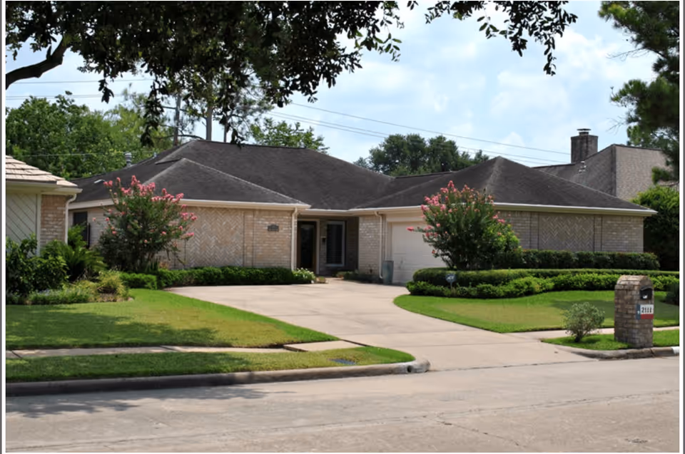 Single-story brick residential building with a dark shingled roof, a driveway leading to a garage, well-maintained green lawn, and flowering bushes in front. The sky is partly cloudy and there are trees framing the top of the image.