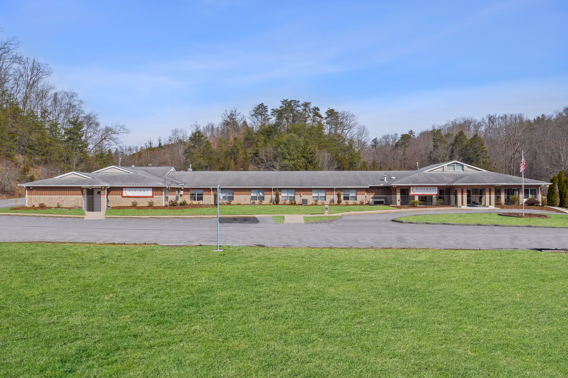 Exterior view of Viviant Healthcare Of Bristol, a single-story brick building with a gray roof, surrounded by a green lawn and trees in the background under a clear blue sky.