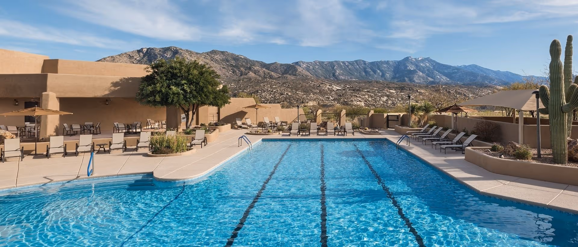 Outdoor swimming pool with clear blue water surrounded by lounge chairs and tables. The pool area is adjacent to a beige building with a shaded patio. In the background, there are desert mountains under a blue sky with some clouds. A large cactus and some desert plants are visible on the right side.