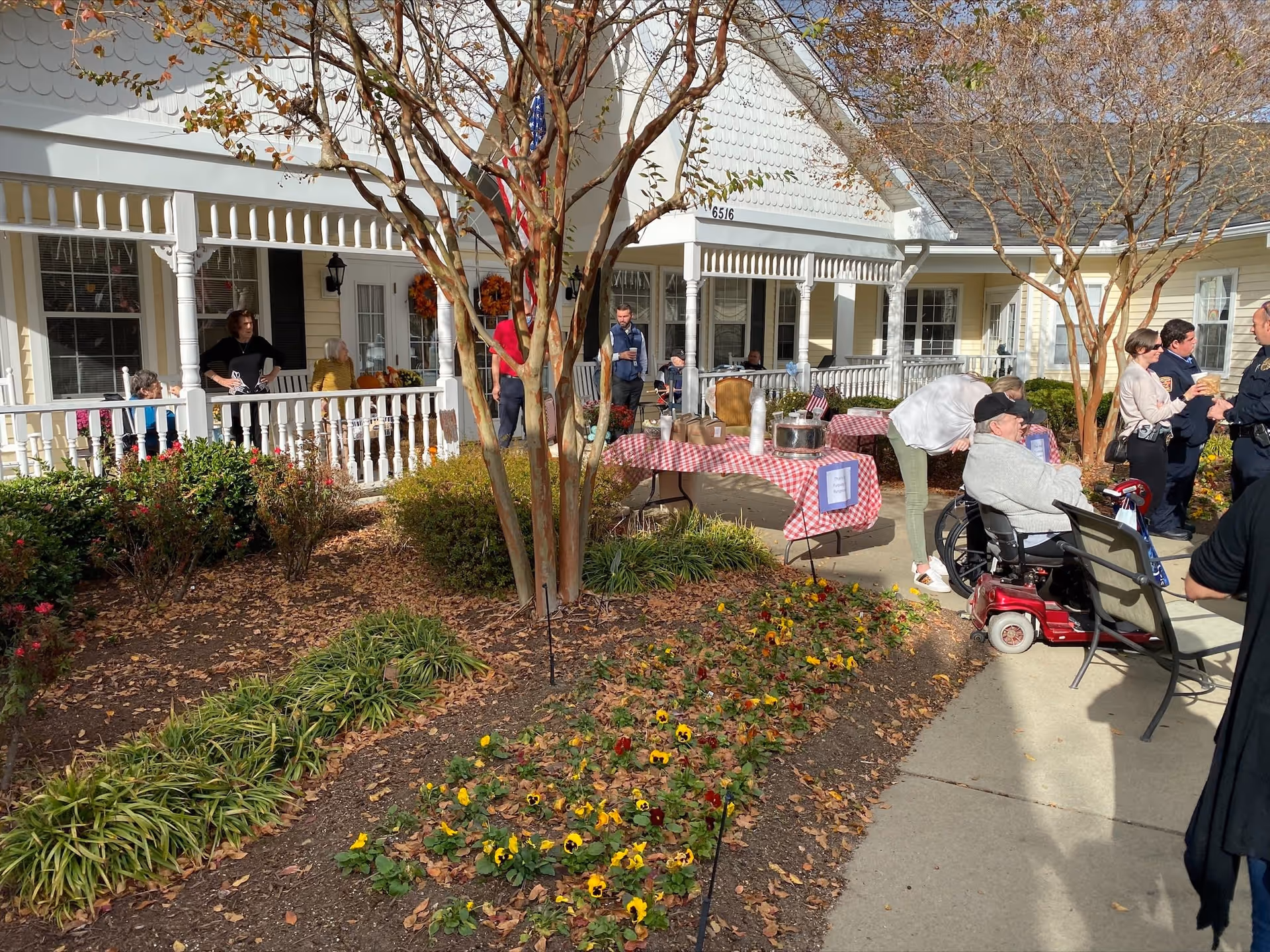 Outdoor scene at TerraBella Fuquay-Varina showing a group of people gathered on a sunny day near a garden with flowers and bushes. Several elderly individuals are seated on benches and in a mobility scooter, while others stand and socialize near a table covered with a red and white checkered tablecloth. The background features a yellow building with white trim and a porch decorated with autumn wreaths.