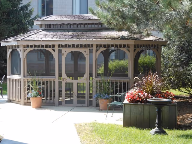 A screened gazebo with a shingled roof situated on a concrete patio surrounded by greenery. There are potted plants with flowers on either side of the gazebo entrance, a green metal chair to the right, and a birdbath in front of a flower planter. Trees and bushes are visible around the area.