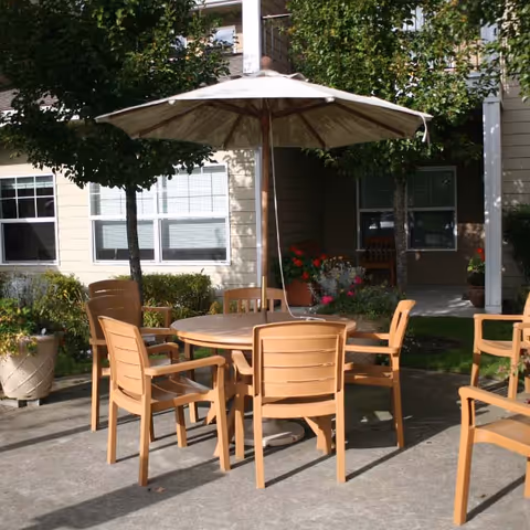 Outdoor patio area with a round wooden table surrounded by six wooden chairs. A large beige umbrella is positioned in the center of the table. The patio is adjacent to a building with windows and surrounded by greenery including trees, bushes, and potted plants with flowers.