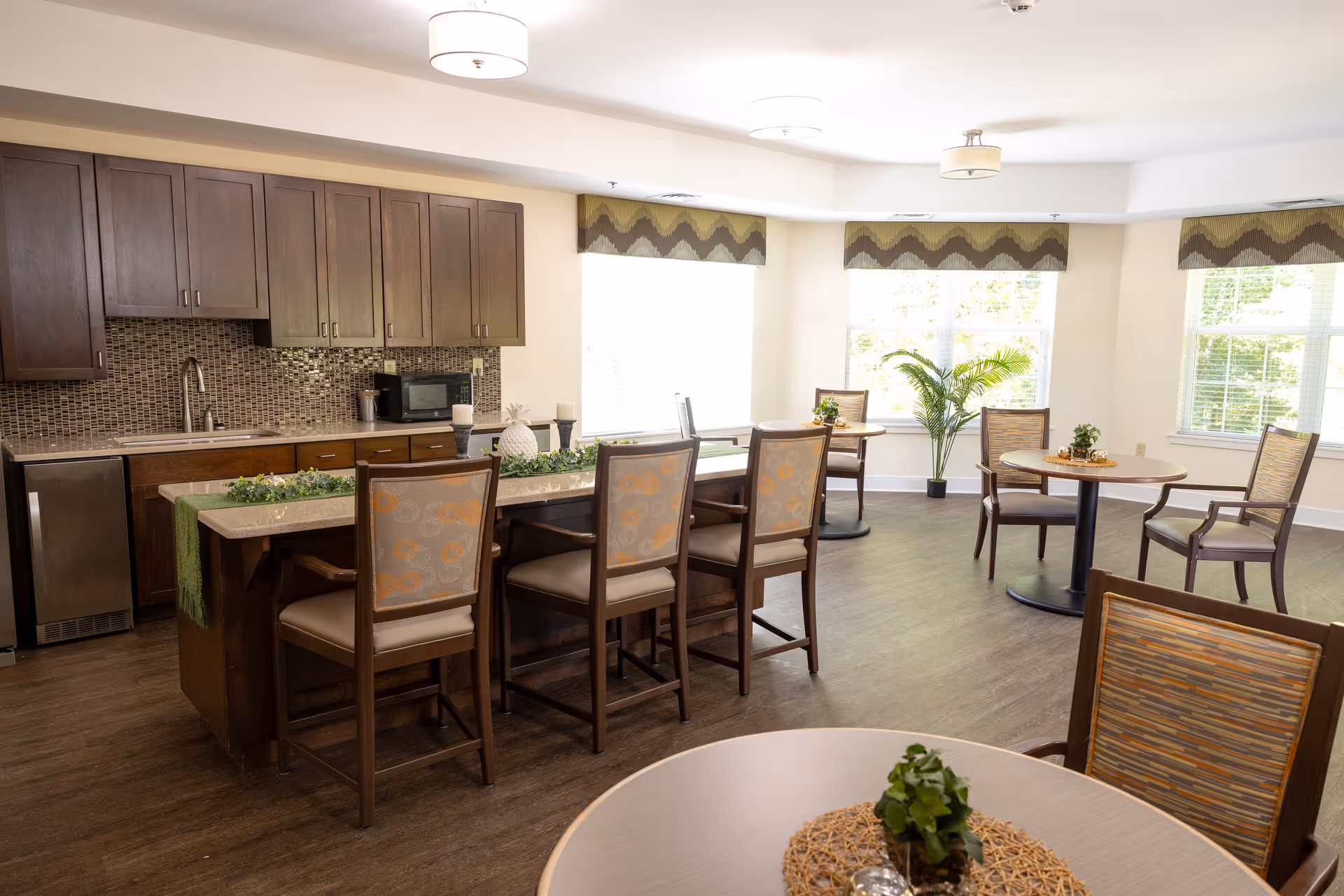 A bright dining area with a kitchen counter featuring four high chairs. The kitchen has dark wooden cabinets, a mosaic tile backsplash, a sink, and a microwave. There are round tables with chairs arranged around the room, some decorated with small plants and placemats. Large windows with patterned valances let in natural light, and a potted plant is placed near the windows.