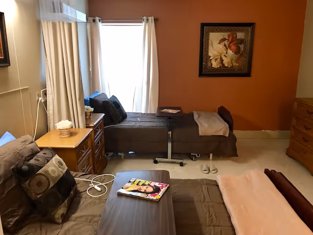 A cozy bedroom in a senior living facility with two single beds, one against a burnt orange wall with a framed floral painting above it, and the other near a window with white curtains. Both beds have dark bedding and pillows. A wooden nightstand with a small flower arrangement and a wooden dresser are visible. A small table with a magazine is placed on one of the beds, and slippers are on the floor near the bed by the wall.