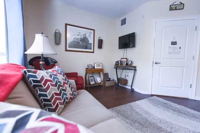 Bright living room with a sofa and patterned pillows, an armchair, wall-mounted TV, framed artwork, and small side tables on hardwood floors.