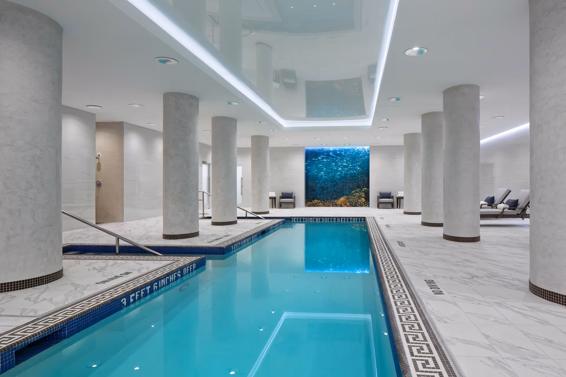 Indoor swimming pool area with clear blue water, surrounded by white marble flooring and large cylindrical columns. There are lounge chairs and small tables along the sides, and a large underwater-themed mural on the far wall. The ceiling has recessed lighting and a reflective panel above the pool.