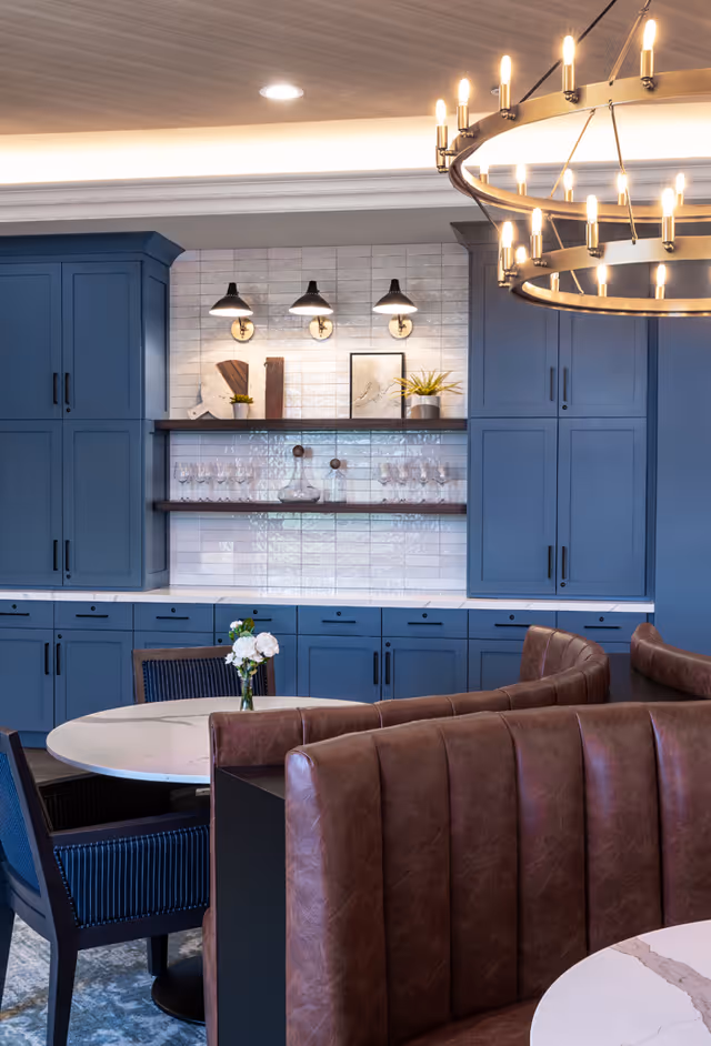 Modern dining area with blue cabinetry, open shelving, round tables, leather banquette seating and a large chandelier.