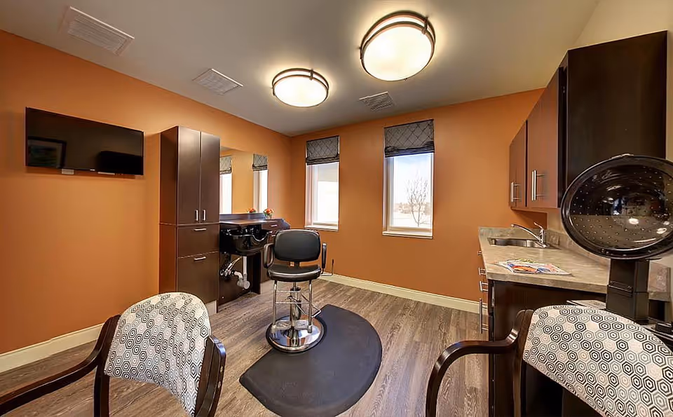 Interior of a senior living facility hair salon with an orange wall, two windows with blinds, a black salon chair on a black mat, a hair washing station, a TV mounted on the wall, two patterned chairs, and cabinetry with a countertop and sink.