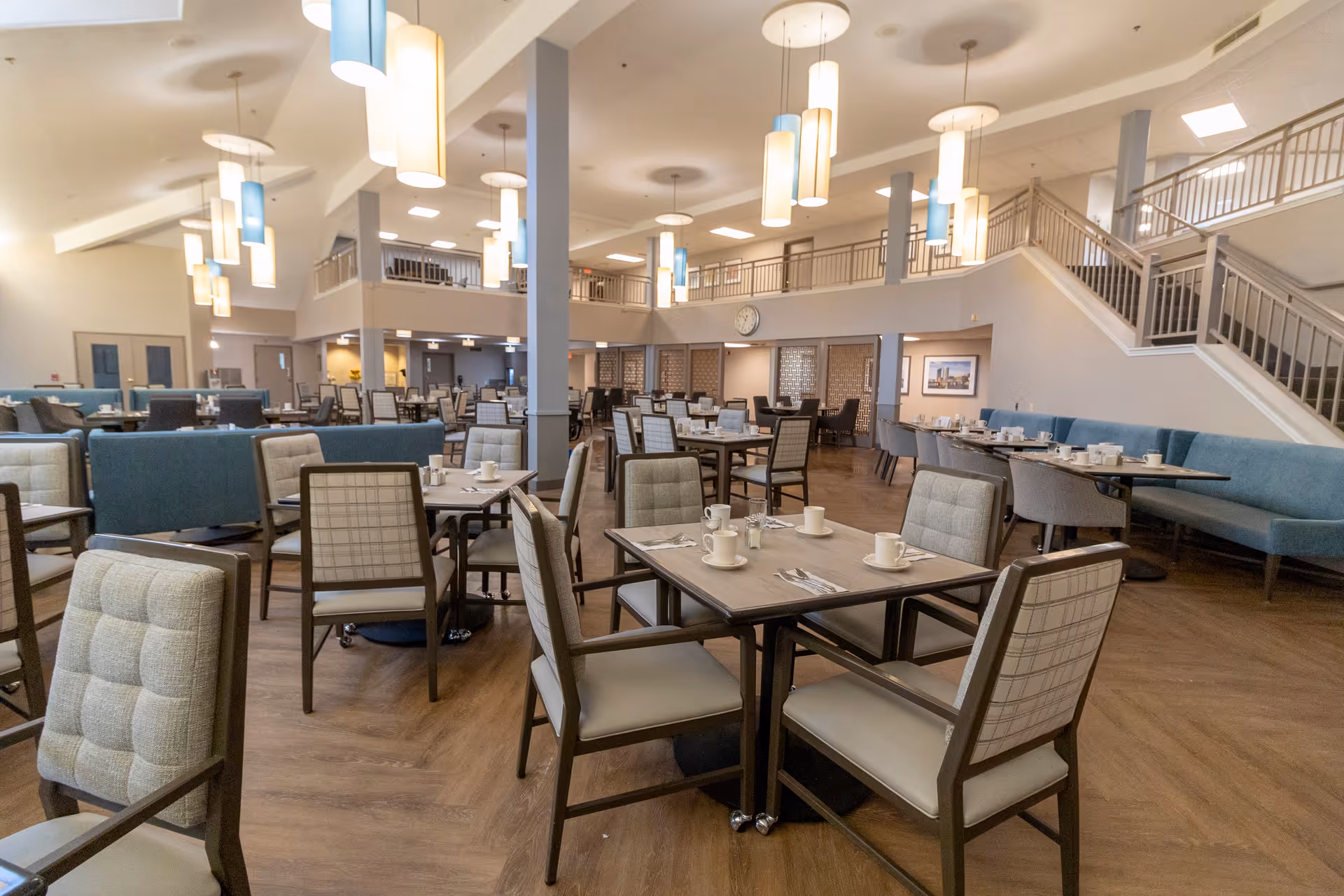 Spacious dining room with multiple tables and chairs arranged neatly. The room features modern pendant lighting, a high ceiling, and a staircase leading to an upper level. Tables are set with cups, saucers, and utensils, and there are blue cushioned benches along the walls.