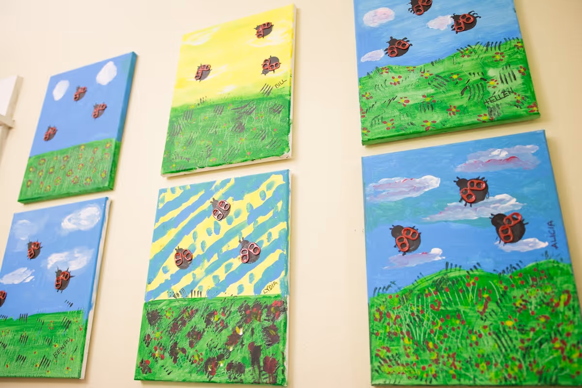 A wall display of six colorful paintings featuring green grassy fields with flowers and blue or yellow skies. Each painting has small red and black ladybug decorations attached, and some paintings have names signed on them.