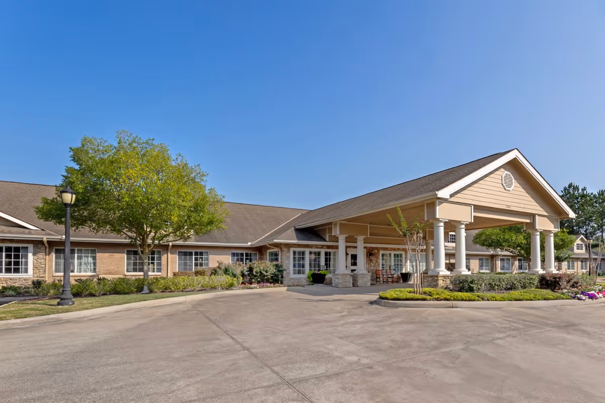 Exterior view of Cinco Ranch Alzheimer's Special Care Center showing a single-story building with a covered entrance supported by white columns, surrounded by landscaped greenery and a clear blue sky.