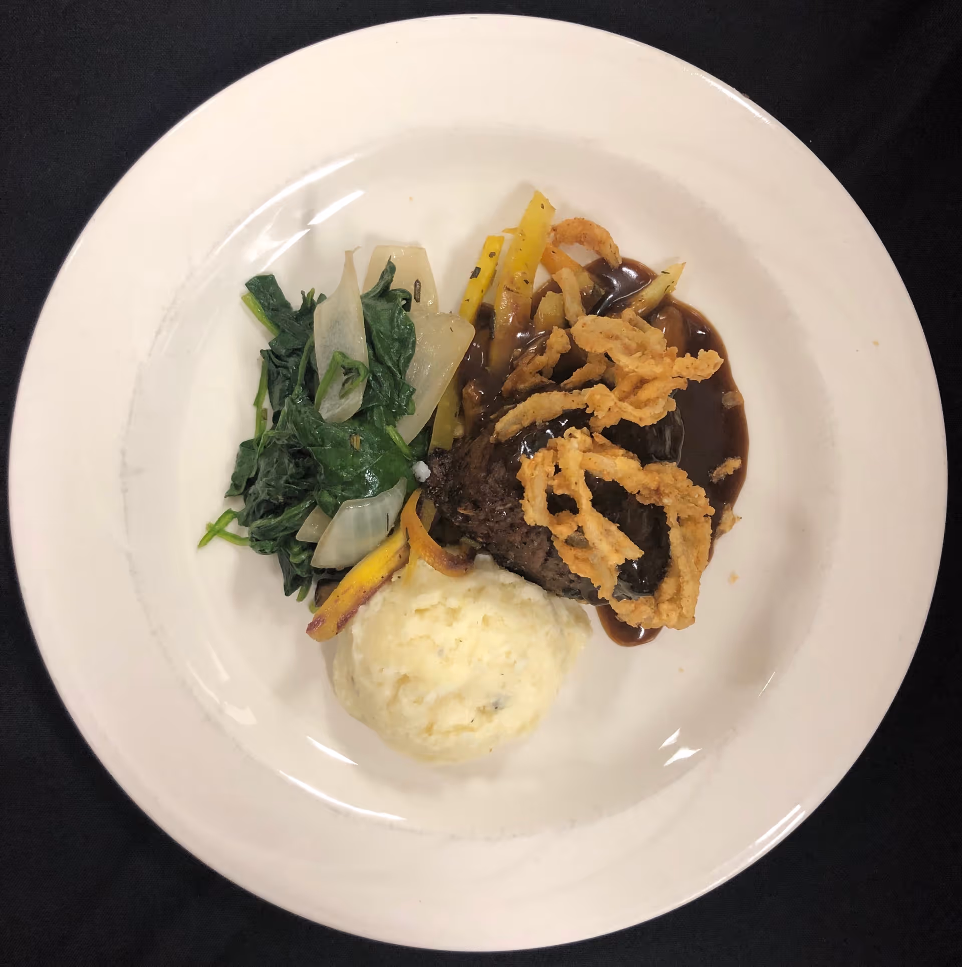 A white plate with a serving of mashed potatoes, sautéed greens with onions, a piece of meat topped with crispy fried onion strings and brown gravy, placed on a black tablecloth.