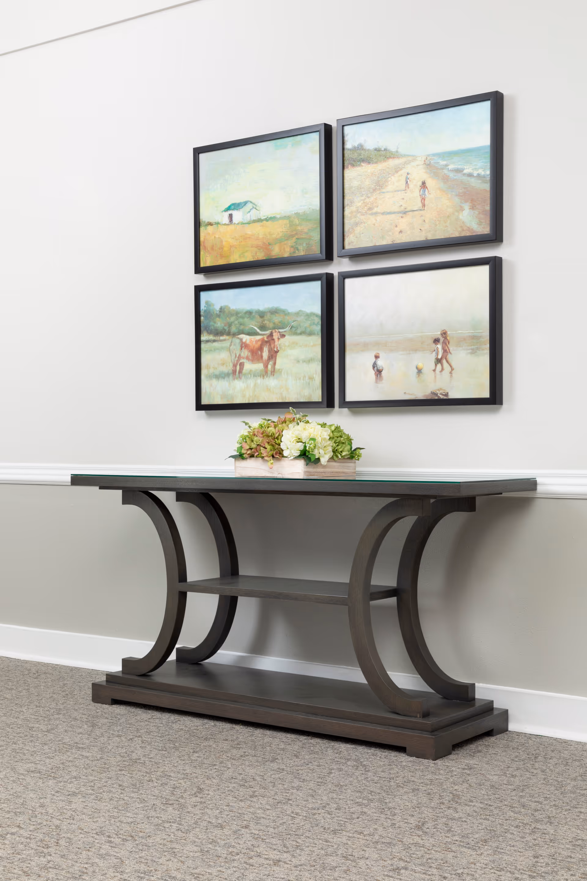 A dark wooden console table with a glass top and curved legs is placed against a light gray wall. On the table is a rectangular planter with green and white flowers. Above the table, four framed paintings are arranged in a square. The paintings depict a small house in a field, a beach scene with two people walking, a cow in a grassy field, and three children playing near water.