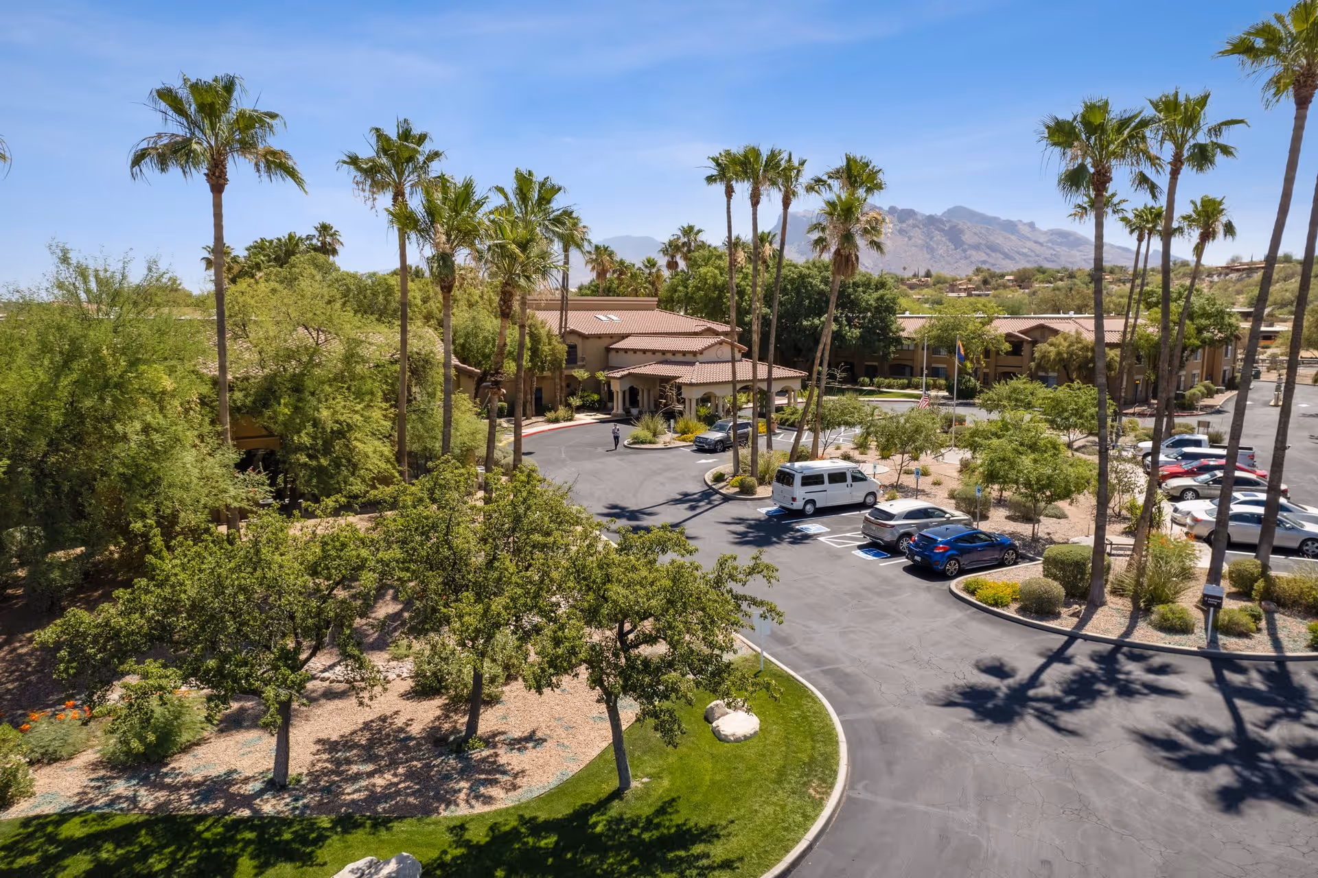 Aerial view of The Country Club of La Cholla facility showing the main building surrounded by palm trees and other greenery, with a parking lot containing several cars and a mountainous landscape in the background under a clear blue sky.