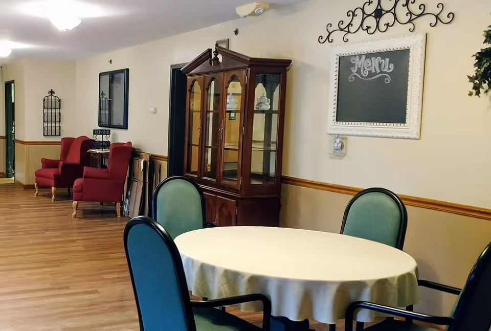 Interior view of a senior living facility room with a round table covered with a white tablecloth and surrounded by four green chairs. In the background, there are two red armchairs, a wooden display cabinet with glass doors, and a wall-mounted chalkboard with the word 'Menu' written on it. The room has wooden flooring and light-colored walls with a wooden chair rail.