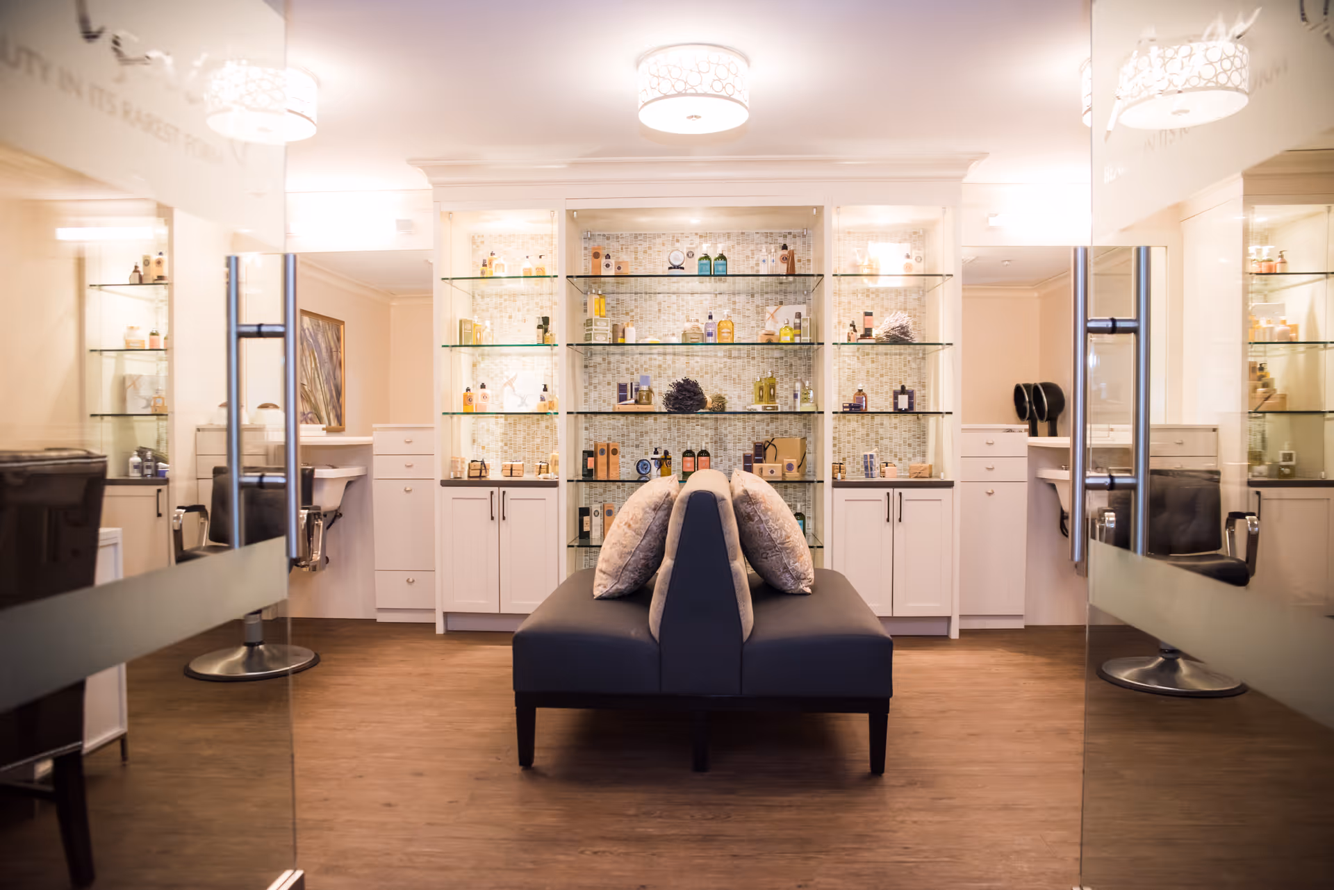 Bright, modern salon-style interior with a central upholstered bench, glass display shelves of products, and styling chairs on either side.