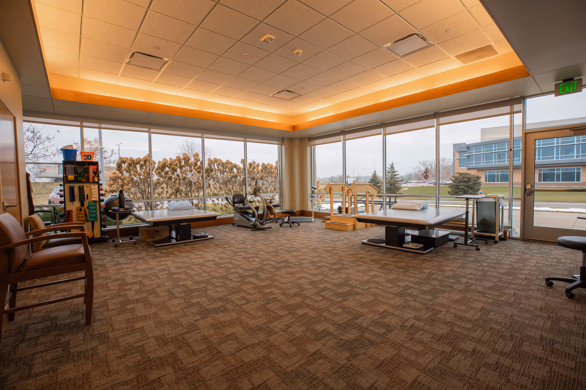 Bright rehabilitation room with therapy tables, exercise equipment and large windows overlooking the grounds.