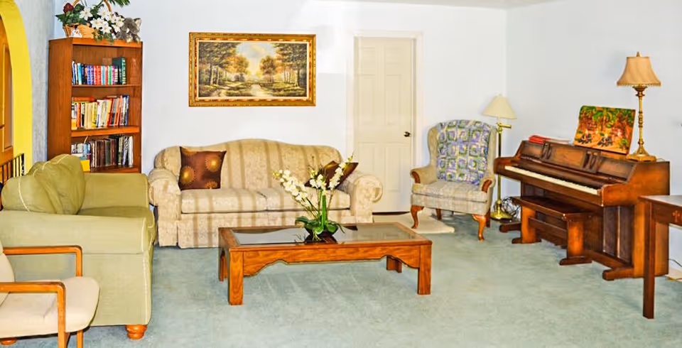 A cozy living room with a beige patterned sofa, a green armchair, a wooden coffee table with a glass top and a flower arrangement, a wooden bookshelf filled with books, a framed landscape painting on the wall, a cushioned armchair with a patterned throw, a wooden piano with a lamp on top, and a side table with a lamp.