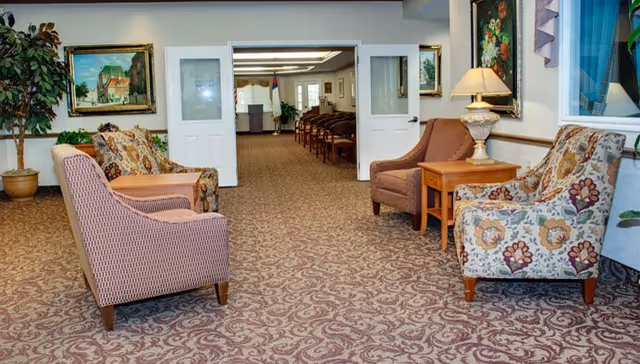 A cozy interior common area with patterned carpet, several upholstered armchairs arranged around wooden side tables, a potted plant, framed paintings on the walls, and a lamp on one of the tables. Double doors lead to another room with rows of chairs and a podium in the background.