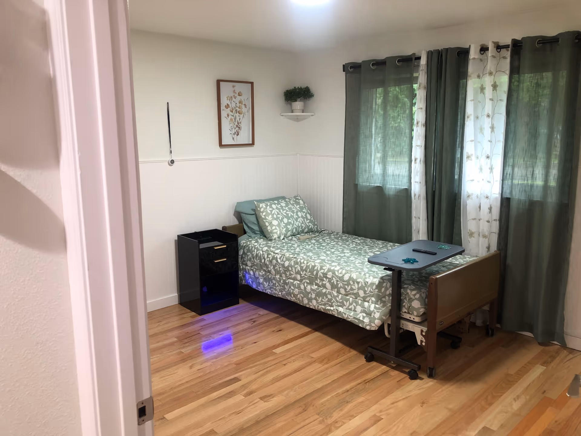 A simple bedroom with a single bed covered in green and white floral bedding. Next to the bed is a black nightstand with two drawers. A small overbed table is positioned over the bed, holding a remote control and two small decorative items. The room has wooden flooring, white walls with wainscoting, a framed floral picture, a small corner shelf with a potted plant, and windows covered with green and white floral curtains.