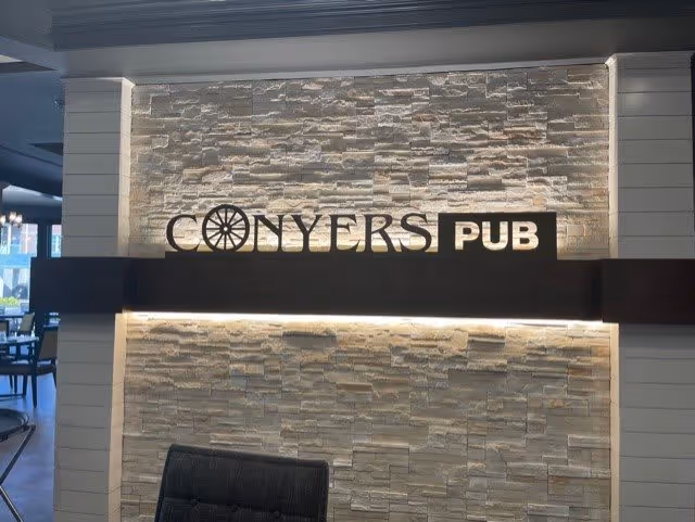 Interior wall with a decorative stone finish featuring a sign that reads 'CONYERS PUB' with a wheel design incorporated in the letter O. Part of a chair and a dining area with tables and chairs are visible to the left.