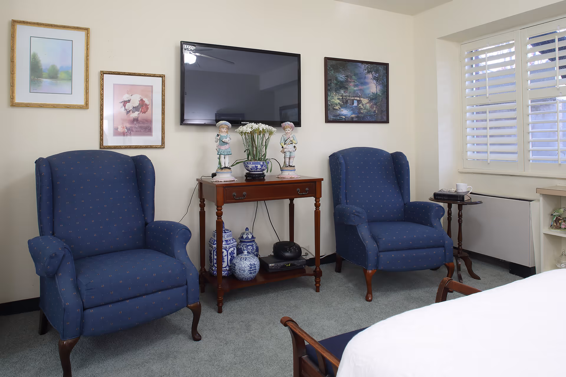 A cozy sitting area in a room with two blue upholstered armchairs facing a wooden table that holds decorative figurines, a vase with flowers, and blue and white ceramic jars underneath. A flat-screen TV is mounted on the wall above the table, and three framed pictures hang on the wall. A window with white plantation shutters is on the right side, next to a small side table with a cup and saucer.