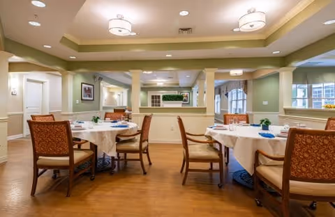 A well-lit dining room with round tables covered in white tablecloths, each set with plates, glasses, and napkins. The room features wooden chairs with patterned upholstery, light green walls, large mirrors, and ceiling lights with circular fixtures. Windows on the right side allow natural light to enter.