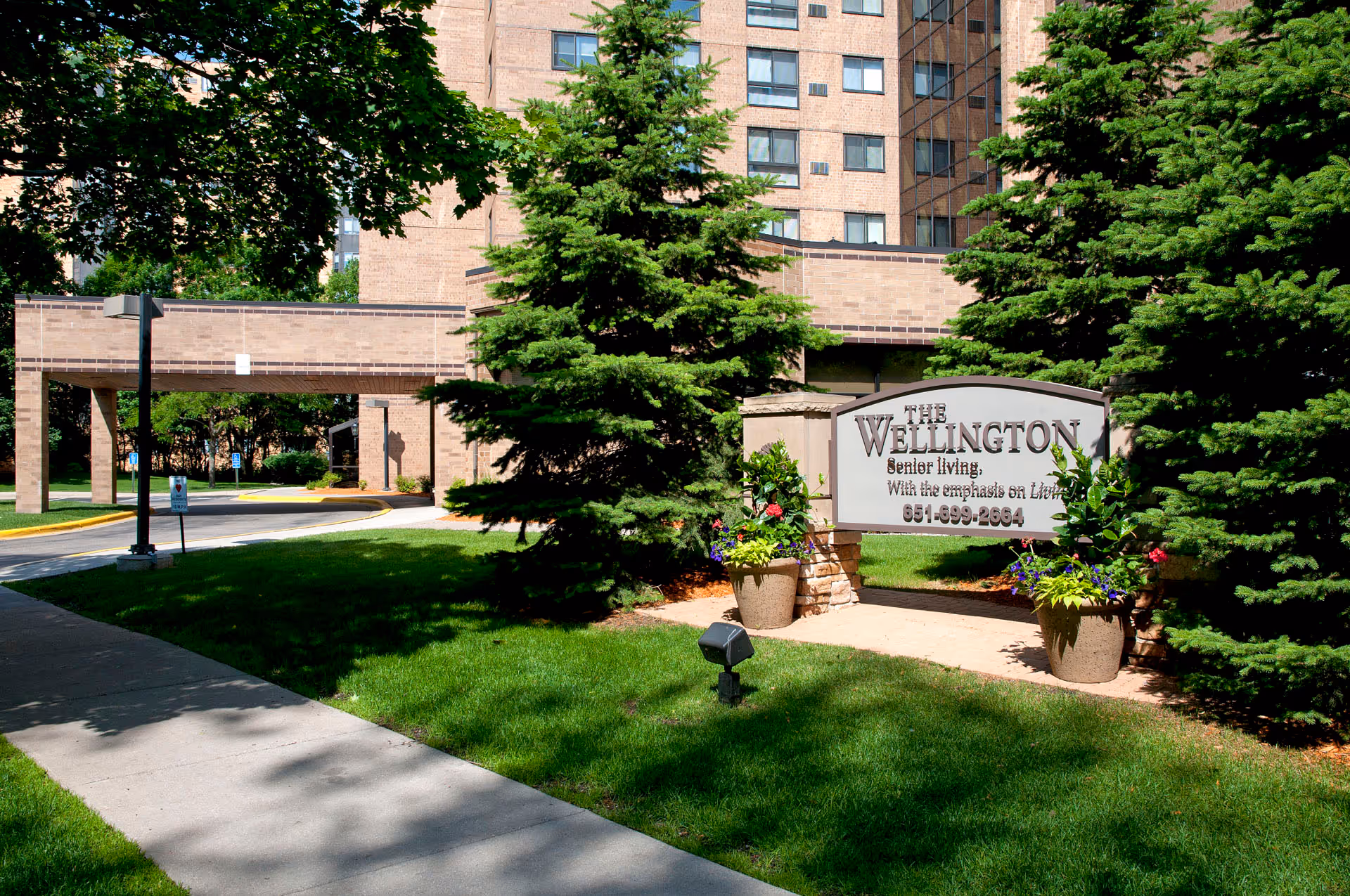 Exterior view of The Wellington Senior Living facility showing a brick building with multiple windows, a covered driveway entrance, green trees, and a sign with the facility's name and phone number surrounded by flower pots.