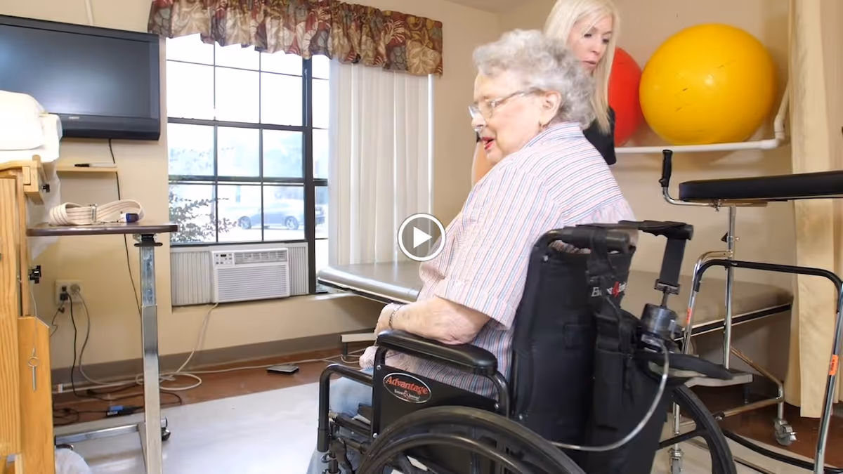 An elderly woman in a wheelchair is assisted by a caregiver in a room with exercise equipment, including large yellow and red therapy balls. The room has a large window with curtains, an air conditioning unit, and a television mounted on the wall.