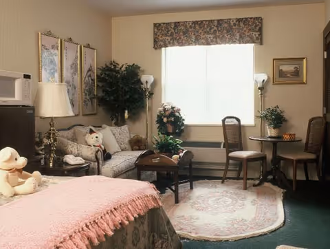 Cozy studio-style bedroom with a bed in the foreground, a sofa and coffee table, a small dining table with chairs, and a window with a floral valance.