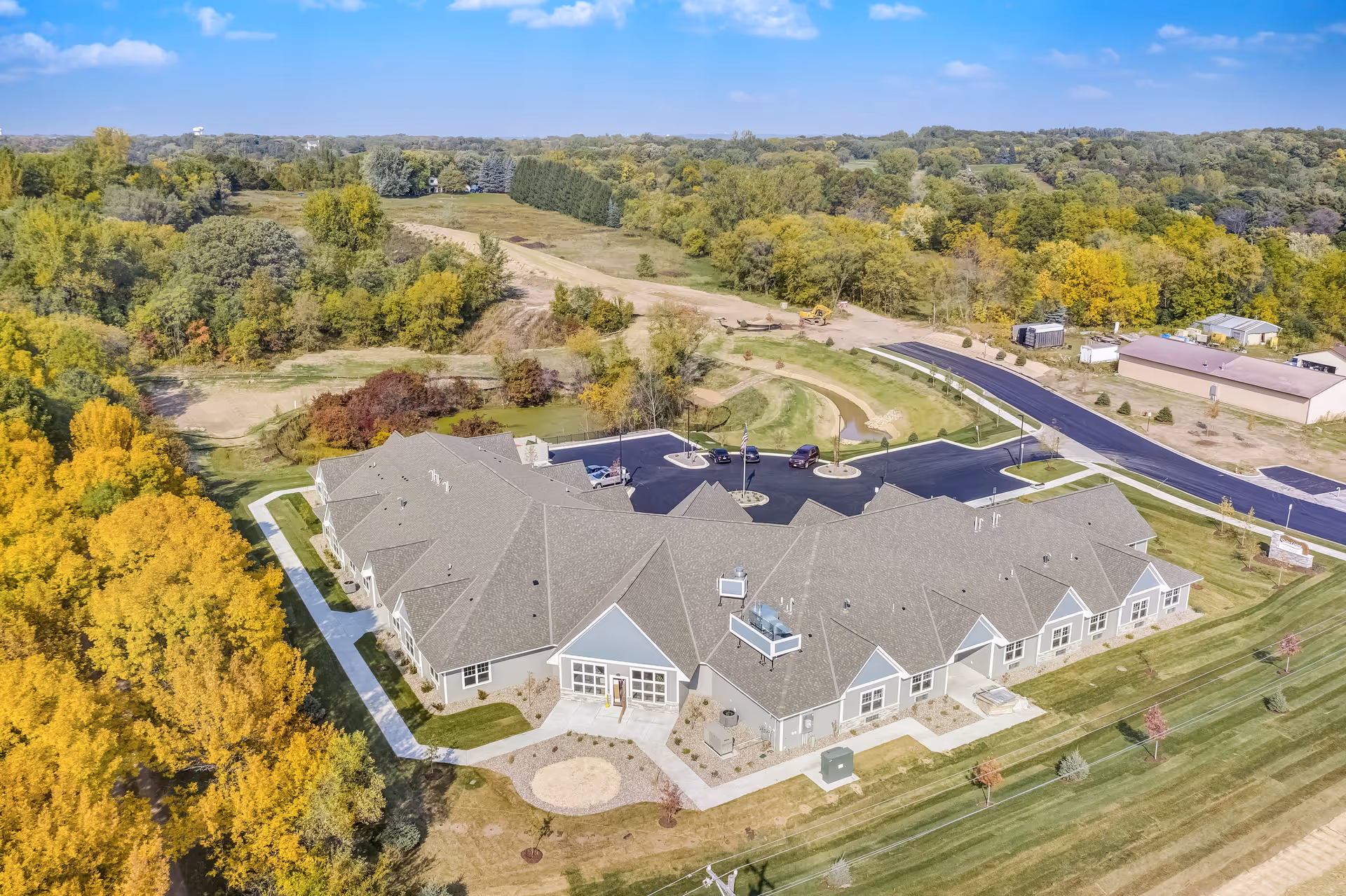 Aerial view of a large single-story assisted living facility surrounded by green lawns and trees with autumn foliage. The building has a gray roof and light-colored exterior walls. There is a paved parking lot with several cars and a curved driveway leading to the entrance. The surrounding area includes open fields and wooded areas under a clear blue sky.