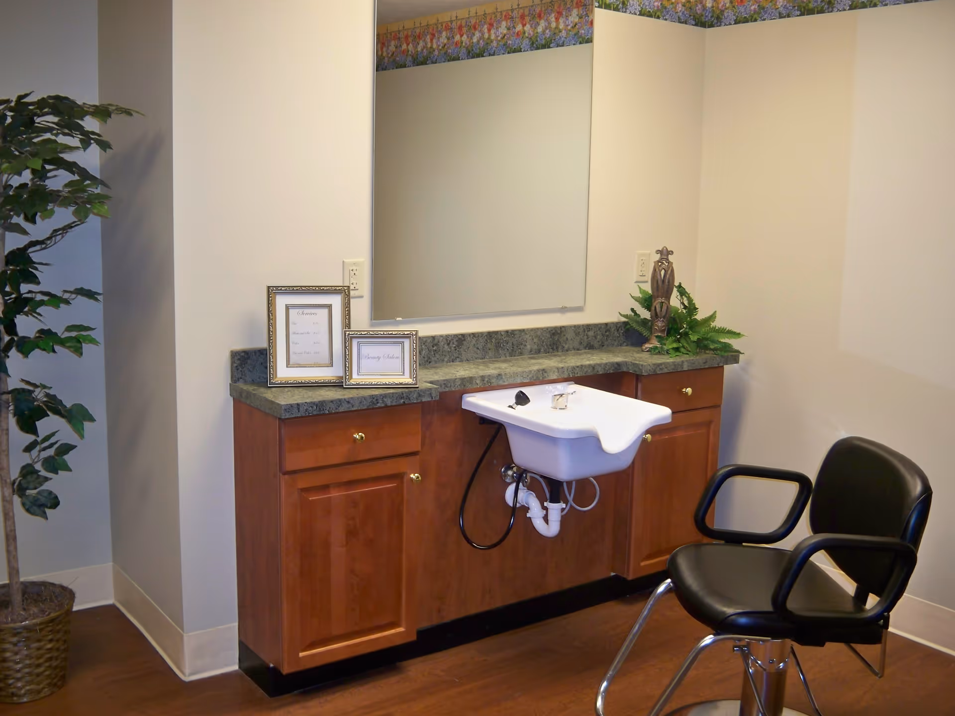 Interior view of a beauty salon area in a retirement community featuring a black salon chair, a white sink with faucet attached to a wooden cabinet with green countertop. There are two framed signs on the countertop and a decorative plant on the right side. A large mirror is mounted on the wall above the sink, and a potted plant is placed on the floor to the left.