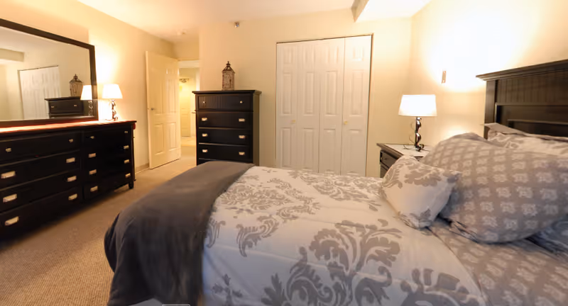 A cozy bedroom in a senior living facility featuring a bed with patterned bedding and multiple pillows, a dark wooden headboard, a nightstand with a lamp, a dresser with a mirror, and a chest of drawers. The room has beige walls and carpeted flooring, with a closet and an open door leading to another area.