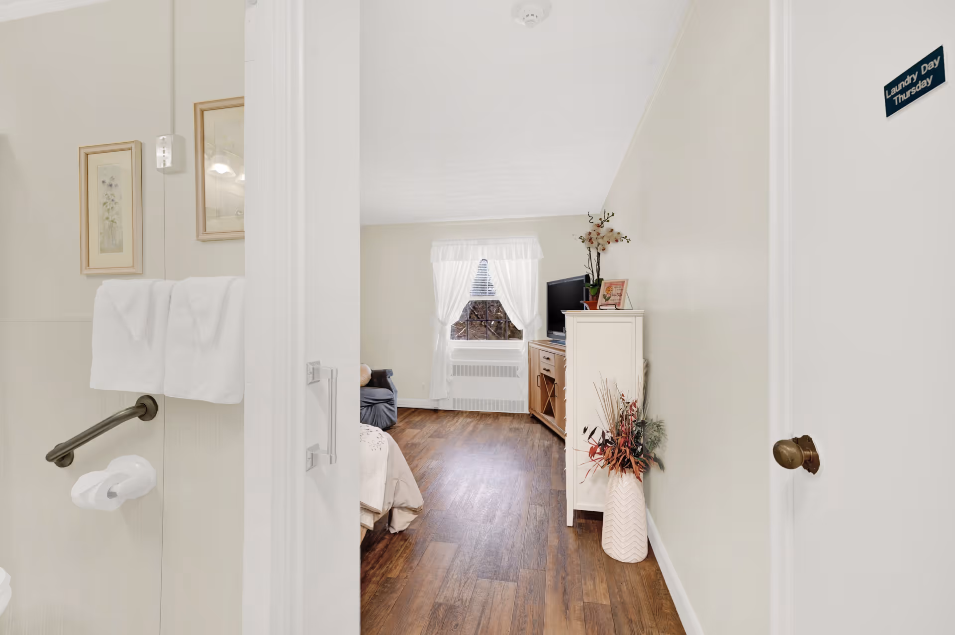 View from a bathroom into a bedroom in a senior living facility. The bathroom has white towels hanging on a rail and framed artwork on the wall. The bedroom features a window with white curtains, a wooden floor, a TV on a cabinet, a small couch, and a vase with decorative plants on the floor.