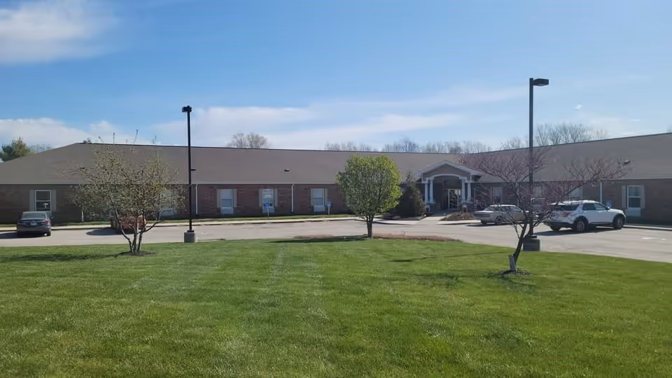Exterior view of a single-story brick building with a pitched roof, surrounded by a green lawn and a few small trees. There are several cars parked in front of the building under a clear blue sky.