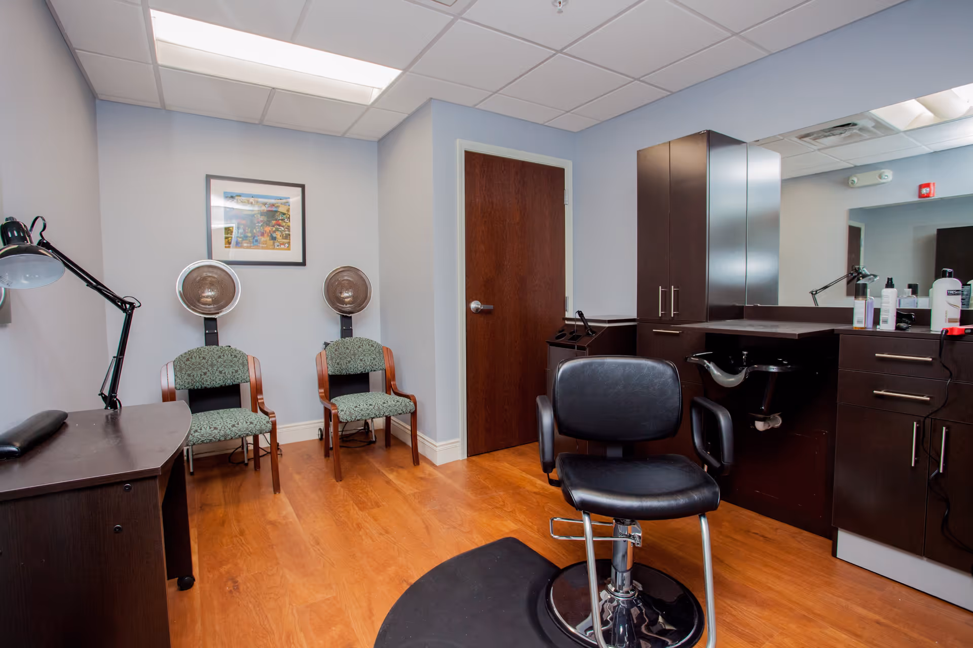 Interior view of a hair salon room with a black salon chair in the center, two green patterned chairs with hair drying hoods against the wall, a wooden door, dark wood cabinetry with a sink, and a large mirror above the counter. The floor is wooden and the walls are painted light blue.
