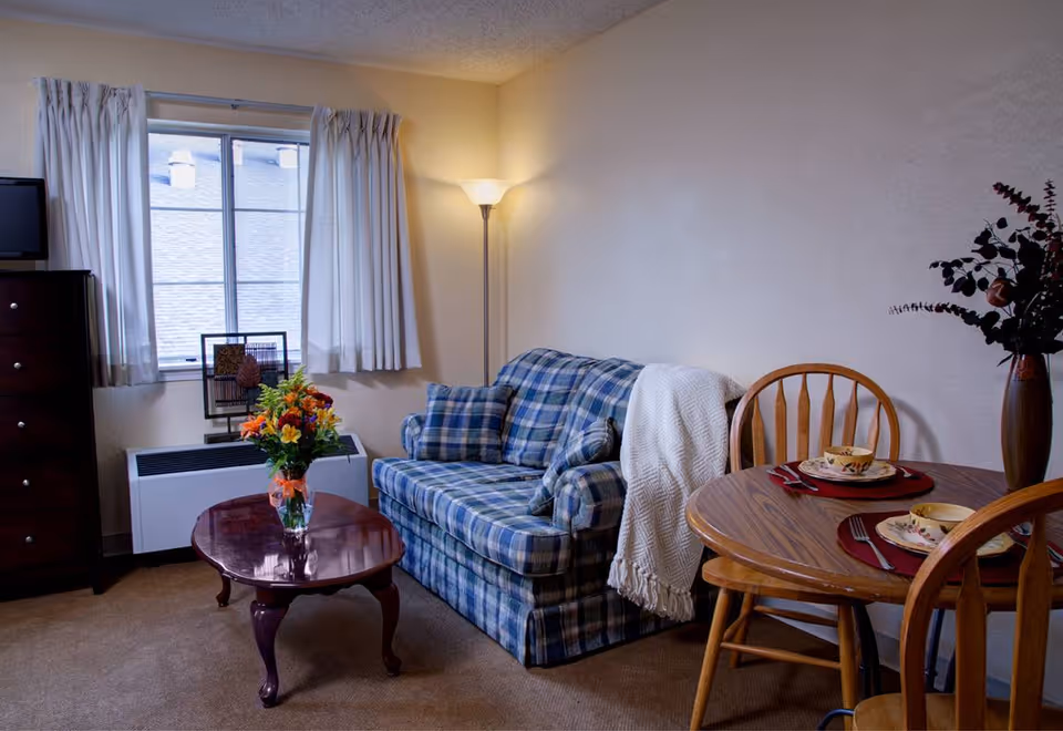 Cozy living room with a plaid sofa, coffee table with a vase of flowers, floor lamp, and a small round dining table set by a window.