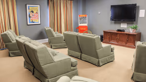 A cozy room with multiple green upholstered armchairs arranged in rows facing a wall-mounted flat screen TV. A wooden cabinet beneath the TV holds electronic devices and a small potted plant. There is a vintage-style popcorn machine in the corner and a framed movie poster on the wall. The room has beige carpet and gray walls with striped curtains covering the windows.