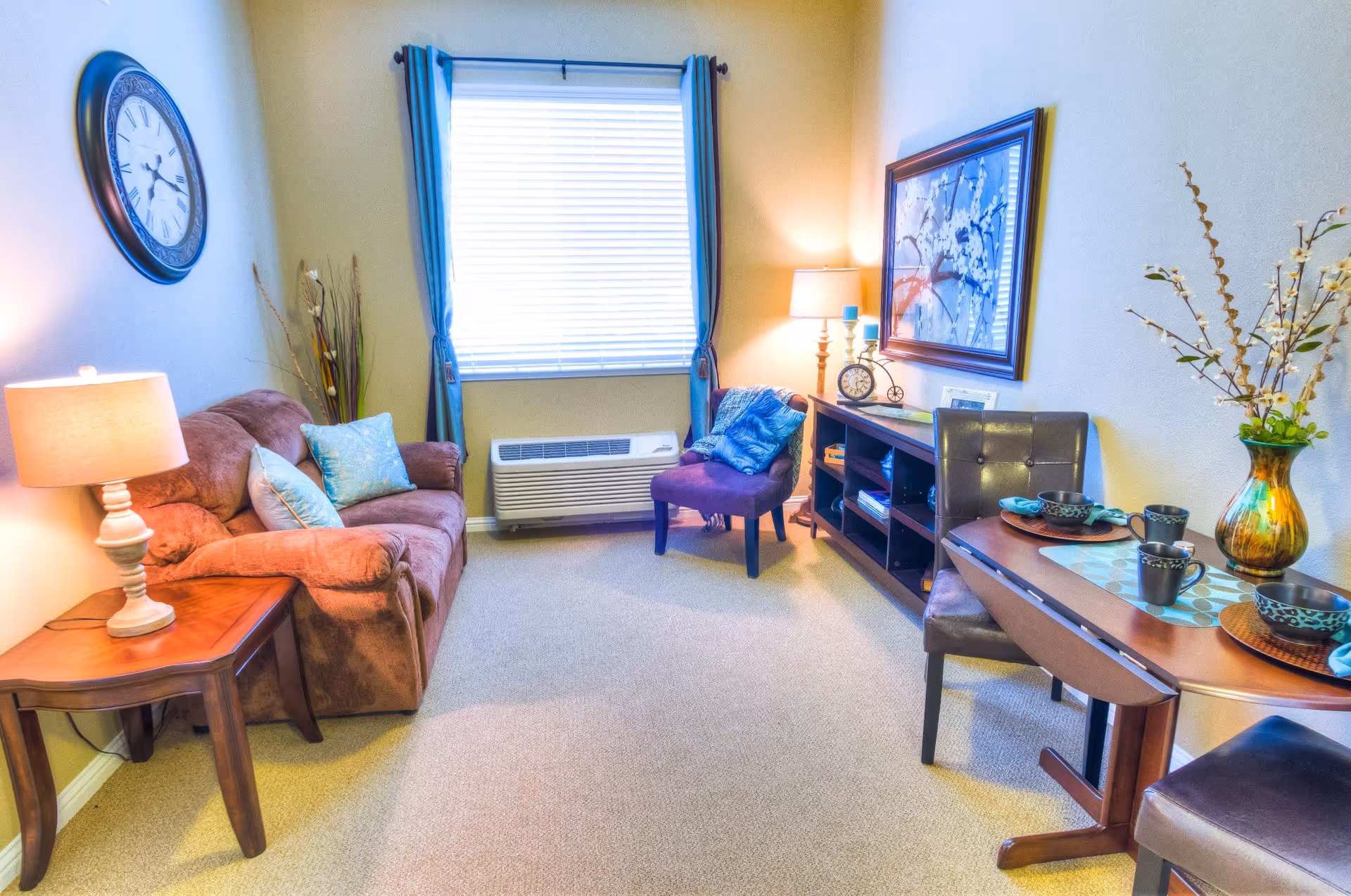 A cozy living room with a brown sofa adorned with light blue pillows, a wooden side table with a lamp, a purple armchair with a blue throw blanket, a wooden console table with decorative items and a framed artwork on the wall. There is a window with blue curtains and a heating/cooling unit beneath it. A small dining table with two chairs is set with bowls, mugs, and a vase with flowers.